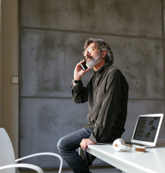 Senior male employee in casual outfit and glasses answering phone call and looking up while sitting on table near laptop in office